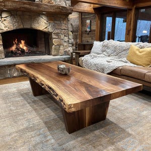 May include: A rectangular coffee table crafted from dark brown walnut wood featuring a natural live edge along the side. The table has a smooth, polished surface and is supported by two solid, thick wooden slab legs that extend to the floor.