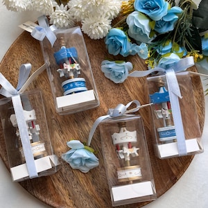 May include: Four miniature carousel figurines in clear boxes, tied with light blue ribbons. The carousel toys are white and blue with gold accents. The background includes blue and white flowers on a wooden tray.