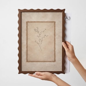 May include: A rectangular wall art piece featuring a dark brown scalloped wooden frame. Inside, a beige fabric panel with frayed edges displays a delicate botanical illustration of a thin plant stem. The frame is sized for A3, 297 by 420 mm.