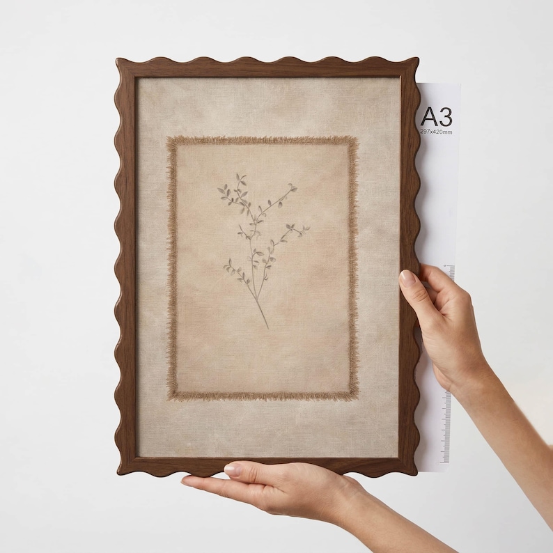 May include: A rectangular wall art piece featuring a dark brown scalloped wooden frame. Inside, a beige fabric panel with frayed edges displays a delicate botanical illustration of a thin plant stem. The frame is sized for A3, 297 by 420 mm.