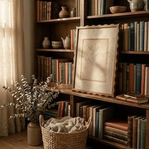 May include: A framed rectangular print featuring a delicate botanical line drawing of a sprig with leaves, centered on a textured beige background. The frame has a wavy, scalloped edge and is positioned on a wooden shelf filled with books.