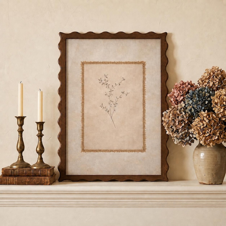 May include: A rectangular wall art piece with a dark brown scalloped frame, featuring a central beige fabric panel with frayed edges and a delicate botanical line drawing of a sprig with small leaves, displayed alongside two brass candlesticks.