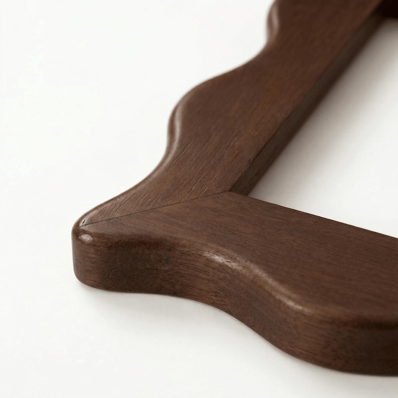 May include: A dark brown wooden frame corner featuring a smooth, wavy, scalloped edge design. The wood grain is visible across the surface of the frame, which has a rounded, soft profile and a mitered joint connecting the two sections of the frame.