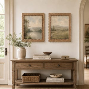 Puede incluir: Dos cuadros rectangulares en marcos de madera festoneados con detalles de cuentas, que muestran pinturas de paisajes con un lago y colinas. El arte se exhibe sobre una mesa consola de madera con dos cajones y un estante inferior.