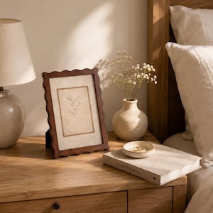 May include: A dark brown wooden picture frame with a wavy, scalloped edge containing a print of a delicate botanical sprig. The frame stands upright next to a small, cream-colored ceramic vase holding white flowers and a small matching dish.