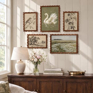 May include: A gallery wall arrangement of five framed prints featuring a swan, a bird, botanical sketches, and landscapes in scalloped wooden frames. Below, a ceramic lamp, a glass vase with flowers, stacked books, and a metal tray sit on a sideboard.