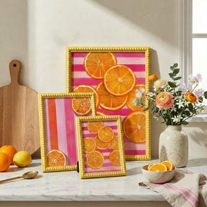 Könnte beinhalten: Vier gerahmte Kunstdrucke mit gelben Perlenrahmen. Die Drucke zeigen Orangenscheiben auf rosa-weiß gestreiften Hintergründen. Eine Vase mit Blumen und eine Schüssel mit Orangenscheiben sind ebenfalls vorhanden, was eine helle und fröhliche Ästhetik schafft.
