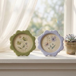 Op de afbeelding: Twee decoratieve fotolijsten met geschulpte randen. De ene is saliegroen, de andere lavendel. Elke lijst bevat een aquarel bloemenprint. De lijsten worden tentoongesteld op een witte vensterbank, met rechts een vetplant in een pot.