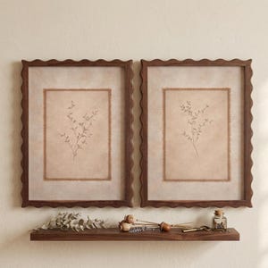 May include: Two rectangular wall frames with dark brown scalloped edges, each containing a beige mat with a frayed edge and a central botanical illustration of a delicate sprig. Below the frames sits a dark wood shelf with dried plant stems.