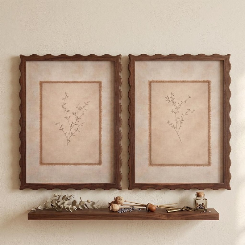 May include: Two rectangular wall frames with dark brown scalloped edges, each containing a beige mat with a frayed edge and a central botanical illustration of a delicate sprig. Below the frames sits a dark wood shelf with dried plant stems.