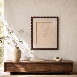 May include: A framed wall art piece featuring a botanical sketch of a sprig with small leaves on a textured beige background, enclosed in a dark brown scalloped frame, displayed above a long wooden media console with a vase and books.