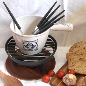 Peut inclure: Un caquelon à fondue en céramique blanche avec un motif floral bleu, doté d'une poignée et d'un support en métal noir avec une grille. Plusieurs fourchettes à fondue aux manches noirs sont à l'intérieur. Une cuillère en bois, des tomates cerises et du pain sont disposés à côté.