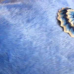 Peut inclure: Une couverture en fausse fourrure bleu clair avec une texture longue et hirsute recouvrant une surface. Un petit coussin rond crocheté multicolore avec des anneaux concentriques crème, bleu clair, bleu foncé et vert est posé sur le côté droit.