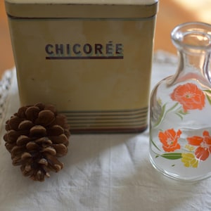May include: A vintage yellow tin with the word "CHICORÉE" printed on the front, a clear glass bottle with orange floral designs, and a brown pine cone rest on a white lace-edged cloth. The items are arranged on a wooden surface.