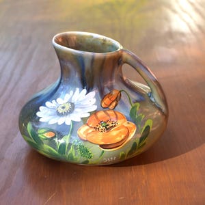 Op de afbeelding: Een keramieken kan met een glanzend, meerkleurig blauw en bruin glazuur. De buik is versierd met handgeschilderde witte madeliefjes en feloranje klaprozen, vergezeld door groene bladeren en stengels op een donkere, gemêleerde achtergrond.