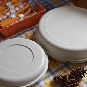 Peut inclure: Deux plats de service en plastique blanc avec couvercles sur une nappe à carreaux. Un panier orange contient des serviettes orange roulées et des ronds de serviette blancs. Plusieurs pommes de pin sont également présentes.