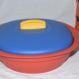 Könnte beinhalten: Ein roter und blauer Lebensmittelbehälter mit gelbem Griff. Der Behälter hat eine runde Form mit blauem Deckel und roter Basis. Der Griff ist rechteckig und gelb. Im Hintergrund befindet sich ein orangefarbenes Gerät.