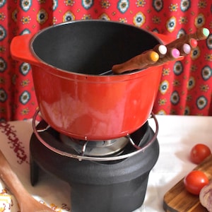 Peut inclure: Une marmite à fondue en fonte émaillée rouge vif avec deux poignées latérales, reposant sur un support en métal noir doté d'une grille circulaire. Une fourchette à fondue au manche marron et aux pointes multicolores est placée dans la marmite.