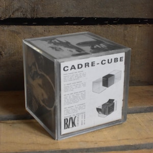 Peut inclure: Un présentoir Cadre-Cube carré et transparent avec un insert imprimé. L'insert présente le texte "CADRE-CUBE" et des schémas. Le boîtier est en plastique transparent et repose sur une surface en bois.