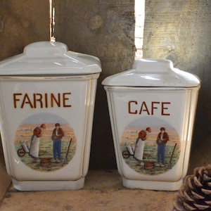 Puede incluir: Dos botes de cerámica blanca con tapas, uno con la inscripción "FARINE" y el otro "CAFE" en letras marrones. Cada bote presenta una ilustración circular de personas en un campo. Estos botes son para la cocina.