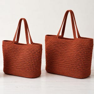 Puede incluir: Tres bolsos tote de ganchillo de color terracota de diferentes tamaños. Cada bolso tiene forma rectangular con base estructurada y dos asas. Los bolsos se exhiben sobre una superficie blanca, mostrando la textura y el color de los artículos hechos a mano.