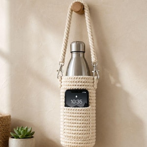 May include: A cream-colored, crocheted water bottle holder with a matching rope strap, holding a stainless steel bottle. A smartphone is in the front pocket, displaying the time 10:35. The holder is suspended from a wooden peg.
