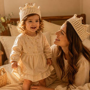 May include: A mother and child are wearing matching cream-coloured crocheted crowns. The child is wearing a cream-coloured dress with small floral accents. The mother is smiling at the child. The scene is set on a bed with cream-coloured bedding.