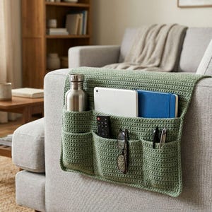 May include: A sage green crocheted sofa armrest organizer featuring multiple pockets. It holds a silver metal water bottle, a white tablet, a blue notebook, a black remote control, a pair of eyeglasses, and two pens in its various compartments.