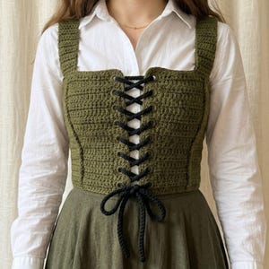Puede incluir: Top de ganchillo verde oliva con detalles de cordones negros. El top presenta un escote cuadrado y tirantes anchos. Los cordones negros recorren verticalmente la parte delantera, creando una apariencia de corsé. Se lleva sobre una camisa blanca con cuello y una falda verde.