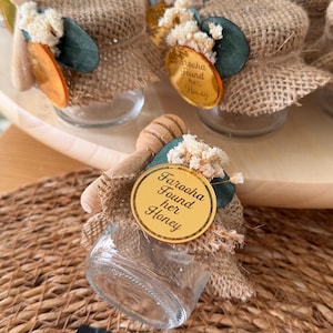Può includere: Piccoli vasetti di vetro pieni di miele, ognuno adornato con un involucro di juta, un cucchiaino di miele in legno e accenti decorativi. Un'etichetta dorata recita "Faraaha Found Her Honey". I vasetti sono disposti su un vassoio di legno, ideali come bomboniera.
