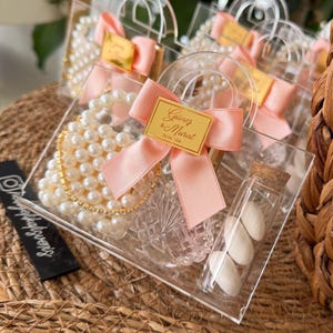Pode incluir: Sacos de presente transparentes de acrílico com colares de pérolas, uma pequena garrafa de vidro com doces brancos e um laço rosa. Cada saco tem uma etiqueta dourada com texto. Os sacos estão sobre uma superfície tecida.