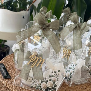 Pode incluir: Caixas de presente de acrílico transparente com laços de fita verde-oliva e detalhes florais. Cada caixa apresenta letras douradas e um arranjo floral decorativo com rosas brancas e vegetação. As caixas estão dispostas sobre uma superfície tecida.