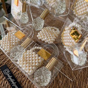 Pode incluir: Frascos de perfume de vidro transparente com tampas douradas s&atilde;o exibidos em sacos de acr&iacute;lico transparente, cada um adornado com enfeites de p&eacute;rolas falsas. Algumas sacolas t&ecirc;m uma etiqueta dourada. Os frascos s&atilde;o dispostos em uma superf&iacute;cie tecida.