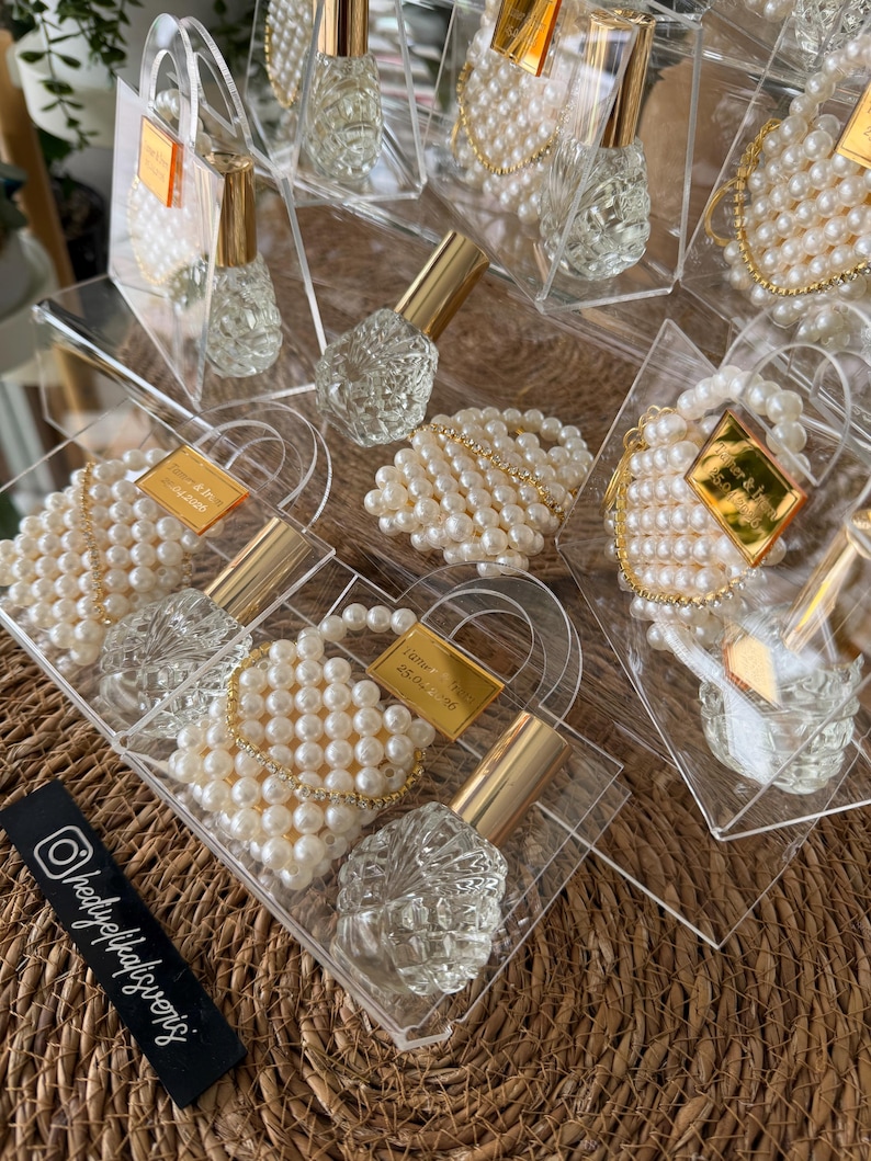 Pode incluir: Frascos de perfume de vidro transparente com tampas douradas s&atilde;o exibidos em sacos de acr&iacute;lico transparente, cada um adornado com enfeites de p&eacute;rolas falsas. Algumas sacolas t&ecirc;m uma etiqueta dourada. Os frascos s&atilde;o dispostos em uma superf&iacute;cie tecida.