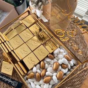 Op de afbeelding: Een goudomrand glazen dienblad gevuld met goud verpakte chocolaatjes, witte en gouden amandelen. De chocolaatjes zijn gegraveerd met initialen en een datum. Een klein boeket en decoratieve gouden letters op de achtergrond.