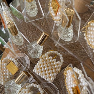 Pode incluir: Frascos de perfume de vidro transparente com tampas douradas s&atilde;o exibidos em caixas de acr&iacute;lico transparente. Algumas caixas cont&ecirc;m pequenas bolsas com contas de p&eacute;rolas e detalhes decorativos com contas de p&eacute;rolas e acabamentos dourados. Os itens est&atilde;o dispostos em uma superf&iacute;cie tecida.