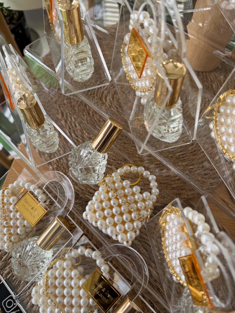 Pode incluir: Frascos de perfume de vidro transparente com tampas douradas s&atilde;o exibidos em caixas de acr&iacute;lico transparente. Algumas caixas cont&ecirc;m pequenas bolsas com contas de p&eacute;rolas e detalhes decorativos com contas de p&eacute;rolas e acabamentos dourados. Os itens est&atilde;o dispostos em uma superf&iacute;cie tecida.