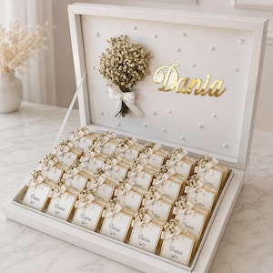 May include: A white gift box filled with chocolates, each topped with a small floral arrangement and a tag reading "Dania" in gold. The box features a gold "Dania" nameplate and is decorated with pearls. The box is open.