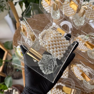 May include: A collection of clear acrylic boxes, each containing a pearl bracelet, a small perfume bottle, and a decorative crystal-cut object. The boxes are arranged on a woven surface, with a hand wearing a black glove holding one.