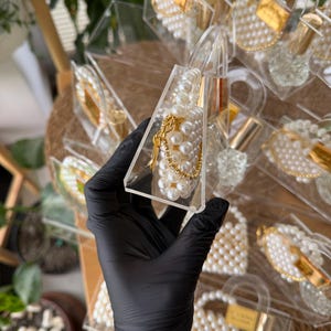 Pode incluir: Lembrancinhas transparentes em forma de bolsa de acr&iacute;lico preenchidas com p&eacute;rolas brancas e detalhes dourados. Cada lembrancinha tem uma corrente dourada e uma pequena etiqueta dourada. As lembrancinhas est&atilde;o dispostas em uma superf&iacute;cie tecida.