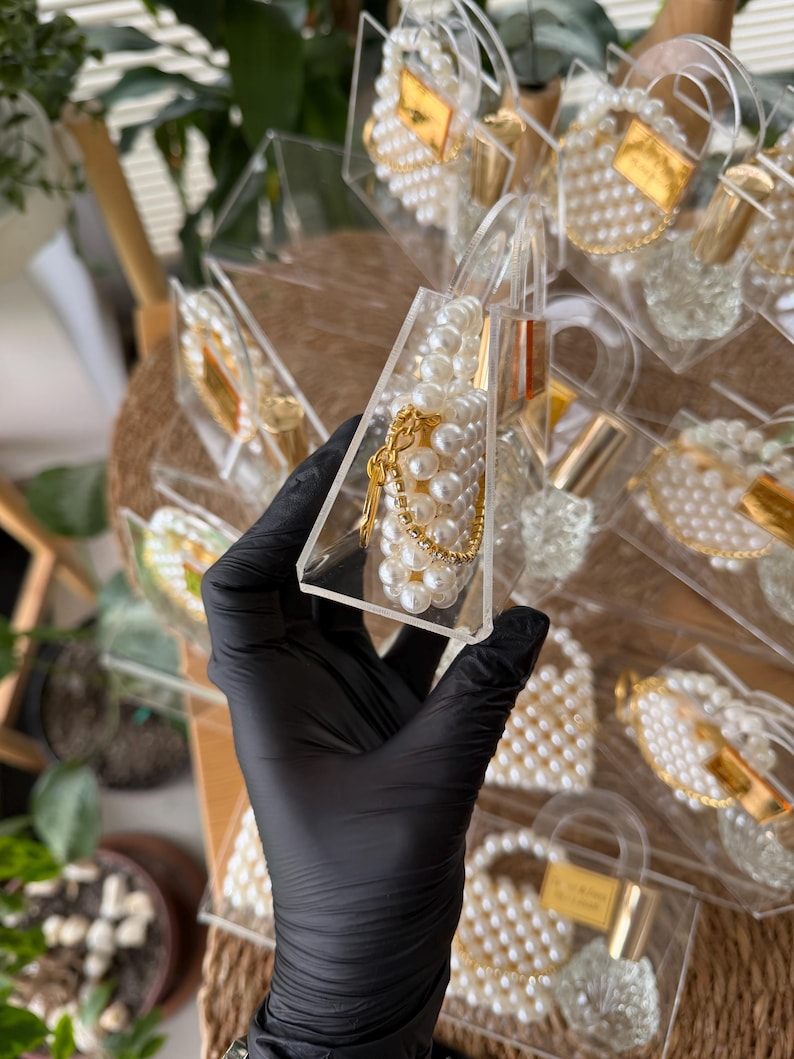 Pode incluir: Lembrancinhas transparentes em forma de bolsa de acr&iacute;lico preenchidas com p&eacute;rolas brancas e detalhes dourados. Cada lembrancinha tem uma corrente dourada e uma pequena etiqueta dourada. As lembrancinhas est&atilde;o dispostas em uma superf&iacute;cie tecida.