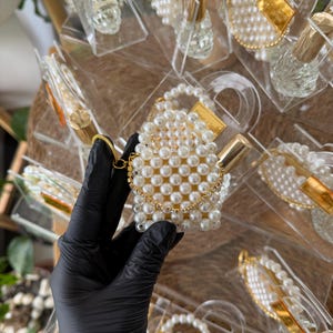 Pode incluir: Uma cole&ccedil;&atilde;o de pequenos frascos de perfume e recipientes em forma de bolsa com contas de p&eacute;rolas. A bolsa tem uma corrente dourada e um fecho dourado. Os frascos t&ecirc;m tampas douradas e s&atilde;o exibidos em suportes de pl&aacute;stico transparente.