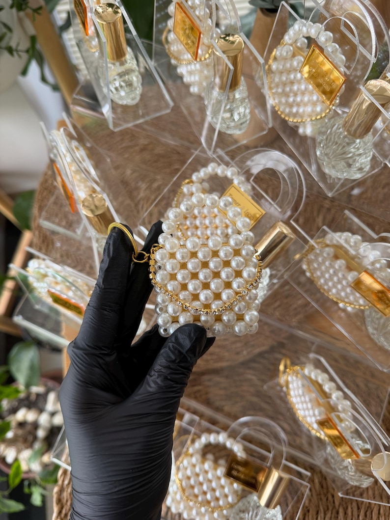 Pode incluir: Uma cole&ccedil;&atilde;o de pequenos frascos de perfume e recipientes em forma de bolsa com contas de p&eacute;rolas. A bolsa tem uma corrente dourada e um fecho dourado. Os frascos t&ecirc;m tampas douradas e s&atilde;o exibidos em suportes de pl&aacute;stico transparente.