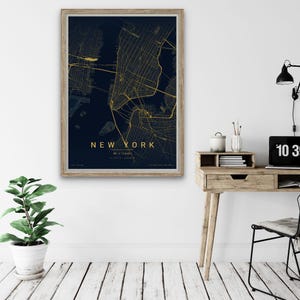 Puede incluir: Un mapa enmarcado de la ciudad de Nueva York, con una cuadrícula dorada de calles sobre un fondo azul oscuro. El nombre de la ciudad está impreso en dorado en la parte inferior. La obra de arte se exhibe en una pared blanca, cerca de un escritorio de madera y una planta en maceta.