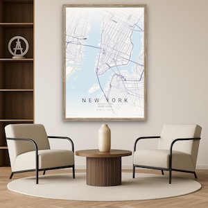 May include: A framed map of New York City in shades of blue and white, with the city's name prominently displayed. The map is in a light wooden frame, hanging above two cream-colored armchairs with black metal frames. A round wooden table and a beige vase are in front of the chairs.