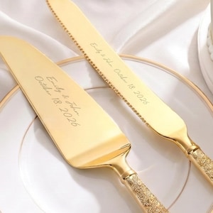 Könnte beinhalten: Ein goldfarbenes Tortenmesser- und Tortenheber-Set mit verzierten, strukturierten Griffen. Die Klingen sind mit dem Text Emily & John October 18, 2026 graviert. Die Utensilien liegen auf einem weißen Teller mit Goldrand auf weißem Stoff.