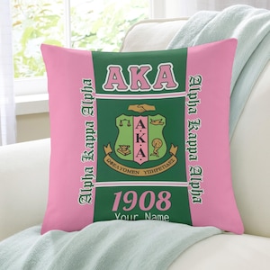 Op de afbeelding: Roze en groene decoratieve kussen met de tekst "AKA" en "Alpha Kappa Alpha". Het kussen heeft een wapen met de Griekse letters AKA en het jaar 1908. De woorden "Your Name" zijn ook zichtbaar.