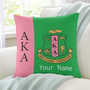 Op de afbeelding: Een decoratief kussen met een roze en groen ontwerp. Het kussen heeft de letters "AKA" in zwart op een roze streep. De groene kant toont een wapen met de letters "AKA" en de tekst "Your Name".