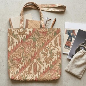 May include: A patterned beige tote bag filled with notebooks, a pen, and glasses. Two art prints and a small drawstring bag are also present. The bag's design incorporates red, green, and beige elements. The bag is ideal for carrying books and stationery.