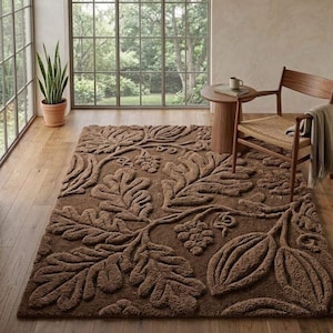 Puede incluir: Una alfombra rectangular marrón con un patrón floral en relieve de hojas y uvas. La alfombra está colocada sobre un suelo de madera, con una silla de madera y una mesa auxiliar en el fondo. También se ve una planta en maceta.